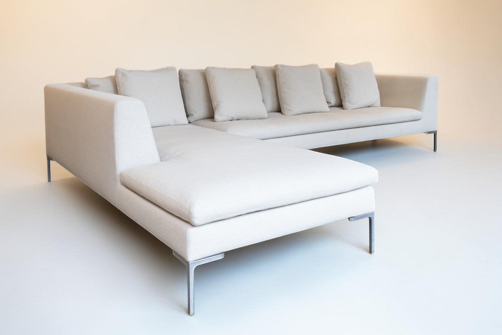 A modern L-shaped sectional sofa upholstered in a light cream fabric with matching back and seat cushions, supported by slim metal legs, set against a minimalist beige background.