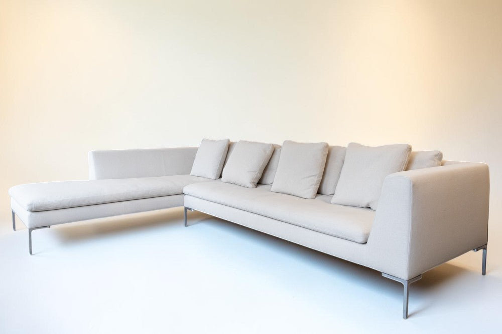 A modern L-shaped sectional sofa upholstered in light beige fabric with matching back cushions and slim metal legs, photographed in a minimalist studio setting with a warm off-white wall and floor.