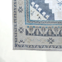 Close-up corner view of a woven rug with a light beige base and blue-gray geometric and floral motifs, shown on a white background.