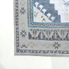 Close-up corner view of a woven rug with a light beige base and blue-gray geometric and floral motifs, shown on a white background.