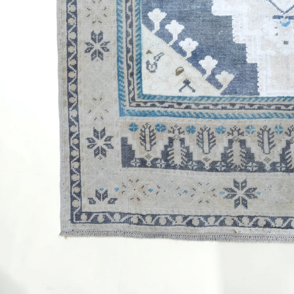 Close-up corner view of a woven rug with a light beige base and blue-gray geometric and floral motifs, shown on a white background.