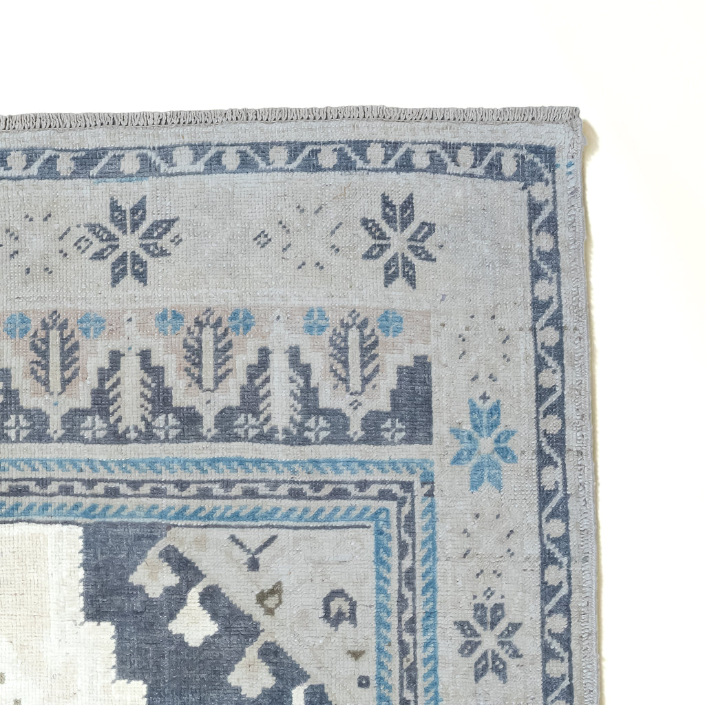 Close-up corner view of a patterned area rug showing a light gray base with blue and dark gray geometric and floral motifs, plus a cream section in the lower left.