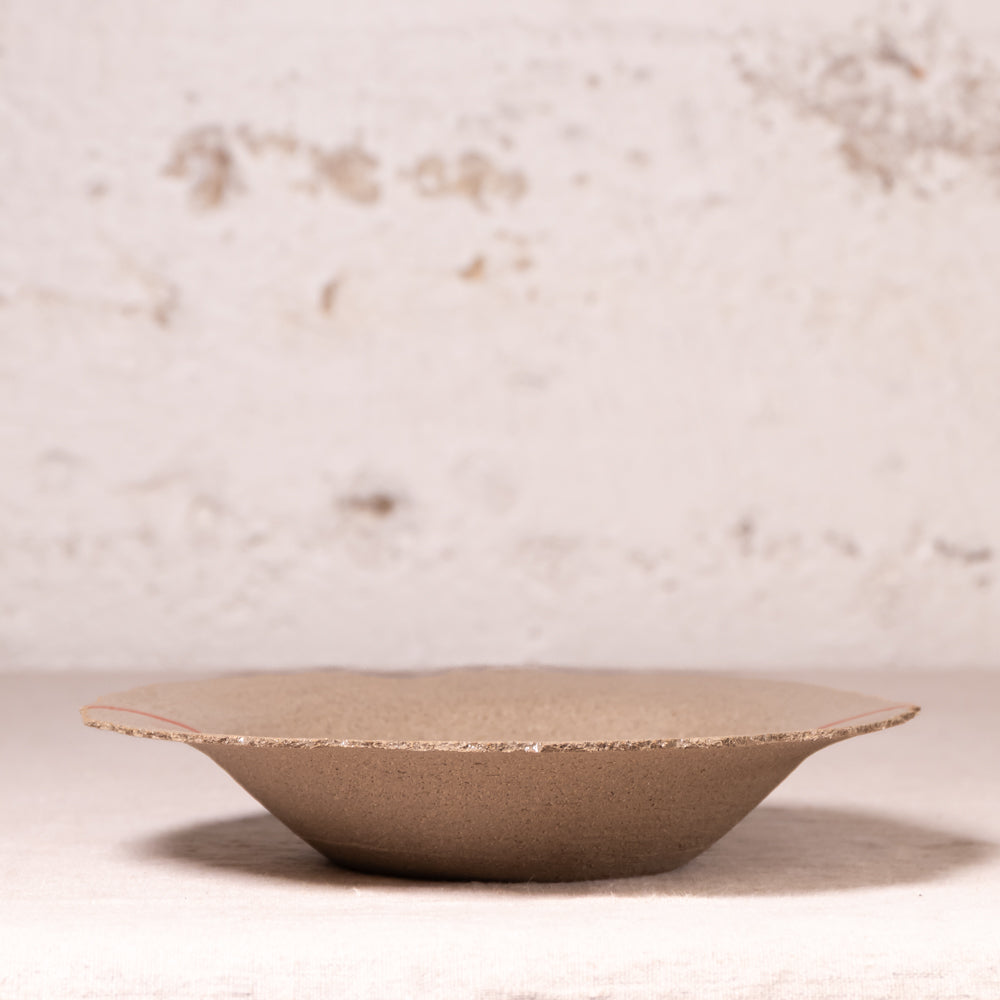 A shallow, wide ceramic plate or dish with a textured, matte taupe-brown finish and a slightly irregular hand-painted edge, set against a rustic, light beige plaster background.