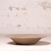 A shallow, wide ceramic plate or dish with a textured, matte taupe-brown finish and a slightly irregular hand-painted edge, set against a rustic, light beige plaster background.