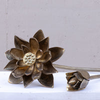 A decorative metal lotus flower sculpture with a textured bronze center and layered petals, shown alongside a smaller bud on a light surface against a neutral gray background.