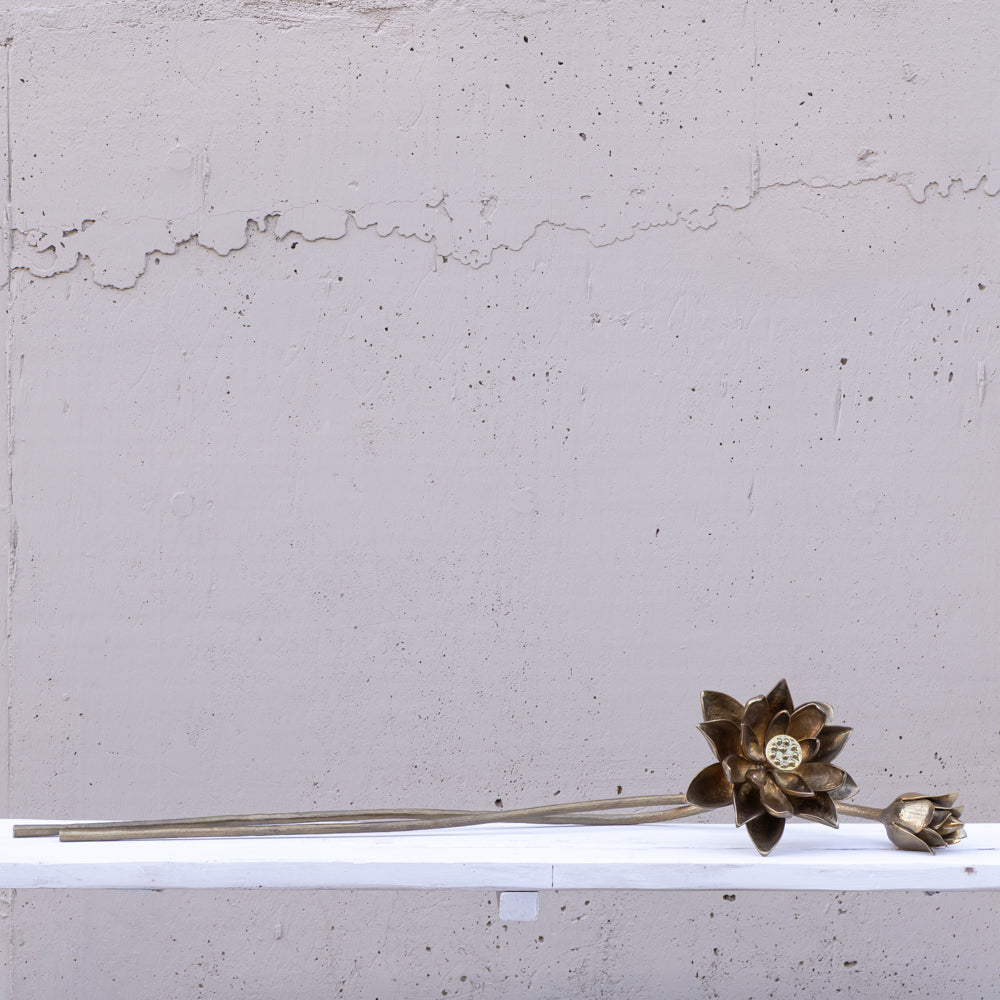 A long, slender decorative object with a metallic bronze lotus flower head and a small bud, resting on a white shelf against a textured light gray wall.