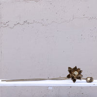 A long, slender decorative object with a metallic bronze lotus flower head and a small bud, resting on a white shelf against a textured light gray wall.