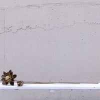 A bronze metal decorative succulent stem with a detailed flower head and a small bud, resting on a white shelf against a textured concrete wall.