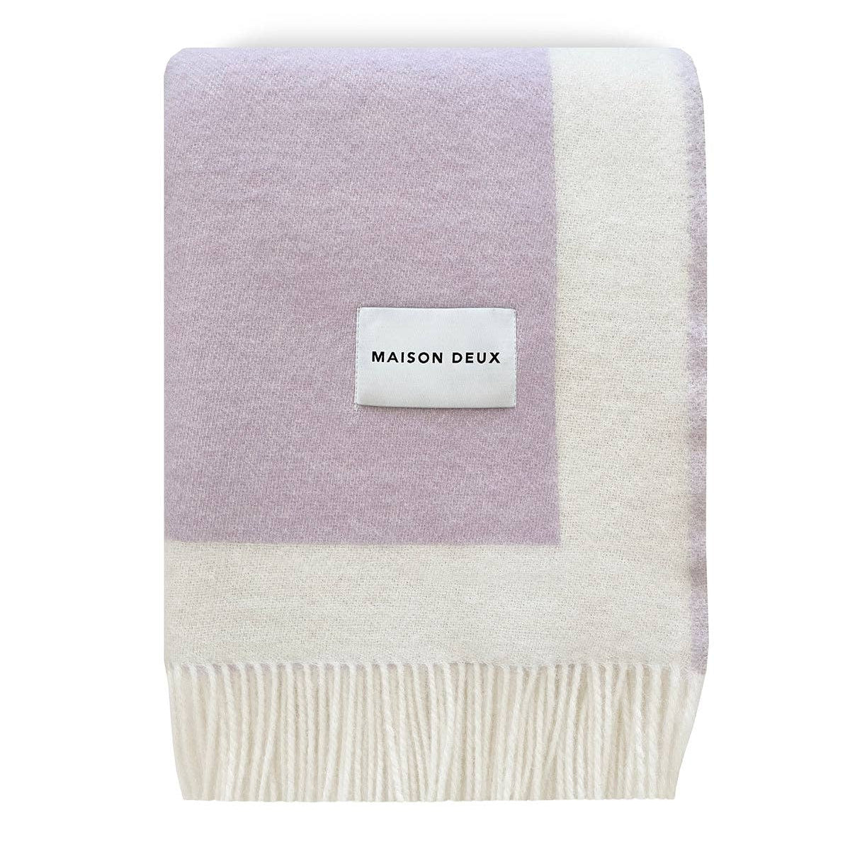 A folded Maison Deux knit throw blanket featuring a lavender purple and off-white color-blocked design with fringed edges and a white brand label.