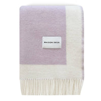 A folded Maison Deux knit throw blanket featuring a lavender purple and off-white color-blocked design with fringed edges and a white brand label.