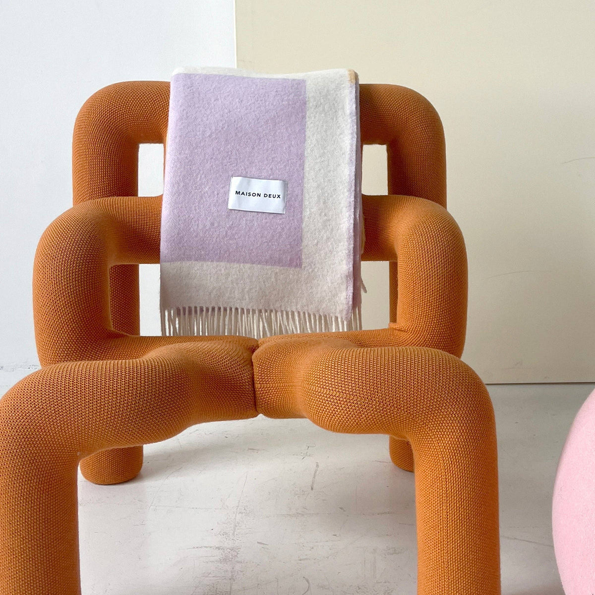 An orange molded armchair with a textured bouclé fabric, styled with a light purple and cream fringed throw blanket that has a visible 'Mossy Oak' label, set against a neutral cream background.