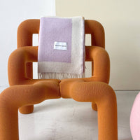 An orange molded armchair with a textured bouclé fabric, styled with a light purple and cream fringed throw blanket that has a visible 'Mossy Oak' label, set against a neutral cream background.