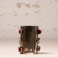 A wide silver cuff bracelet featuring four red faceted gemstones set in silver bezel settings, displayed against a textured off-white background.