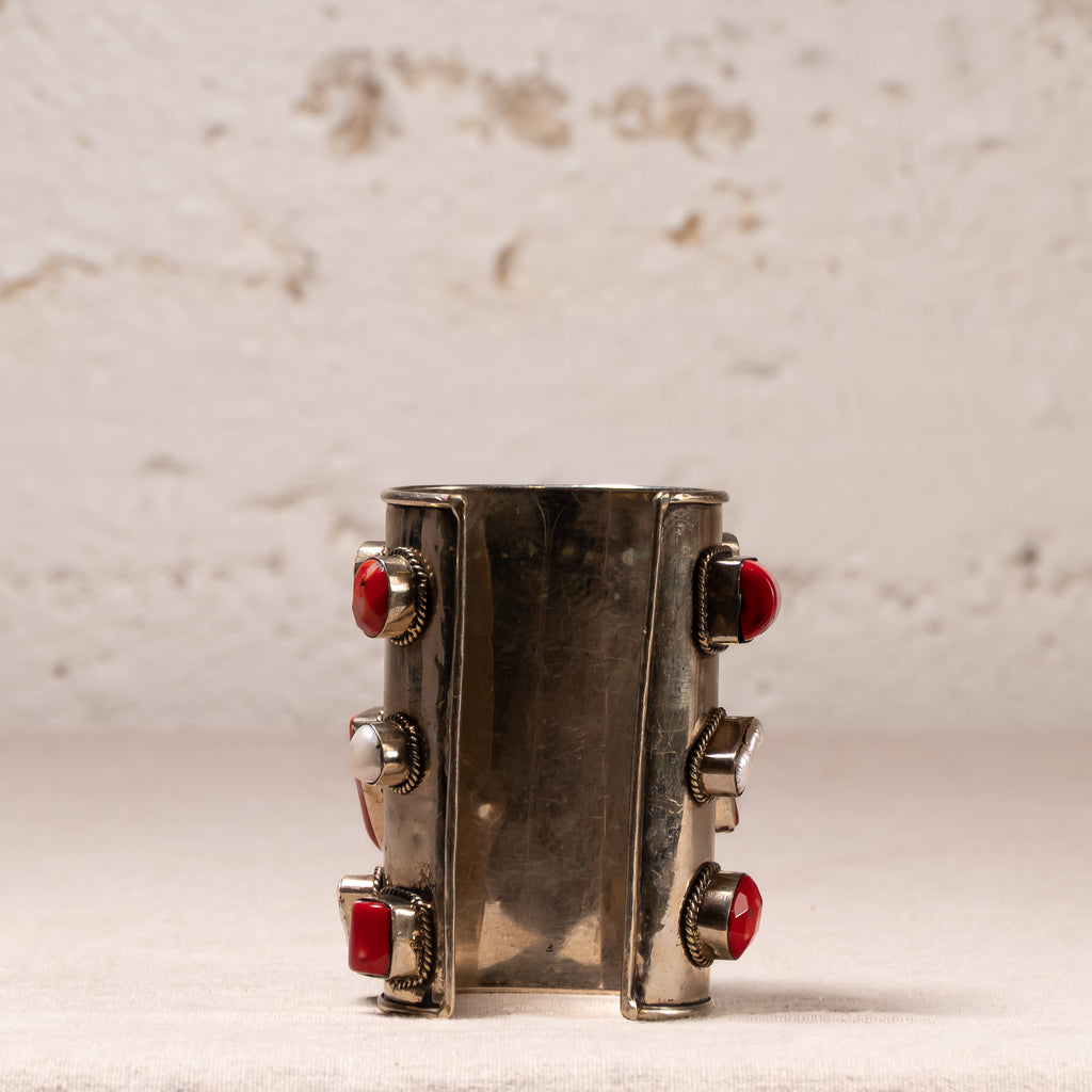 A wide silver cuff bracelet featuring four red faceted gemstones set in silver bezel settings, displayed against a textured off-white background.