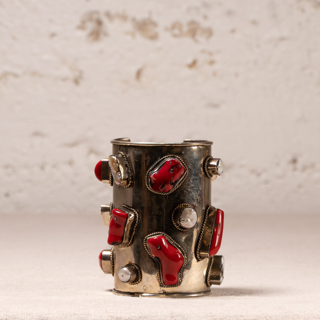 A vintage silver cuff bracelet decorated with red coral inlays and small white pearl accents, displayed against a textured beige background.