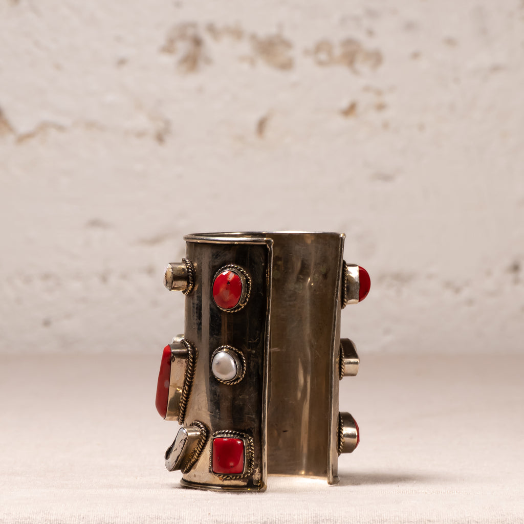 A wide silver cuff bracelet decorated with red gemstones and a central white pearl, displayed on a light textured surface against a neutral background.