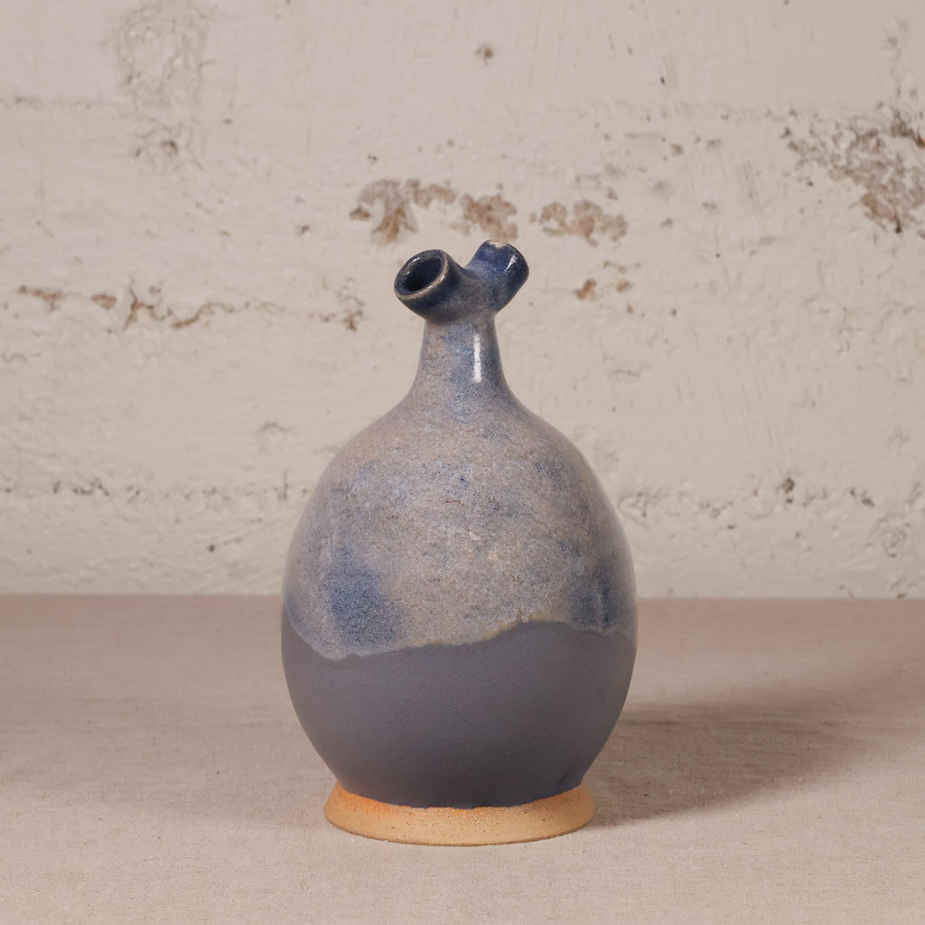 A handmade ceramic vase with a rounded bulbous body, a short flared neck, and a small side spout, featuring a blue and gray glaze over a natural clay base, set against a rustic textured wall.