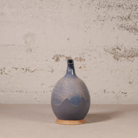 A handmade ceramic vase with a bulbous body and a short, narrow neck, featuring a blue and gray glaze with a natural terracotta base, set against a textured off-white wall.