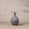 A handmade ceramic vase with a bulbous body and a short, narrow neck, featuring a blue and gray glaze with a natural terracotta base, set against a textured off-white wall.