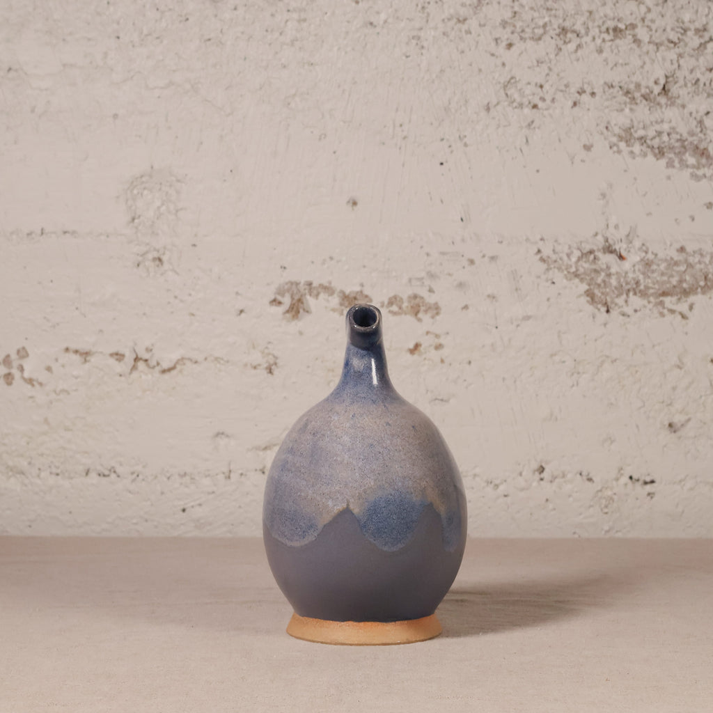 A handmade ceramic vase with a bulbous body and a short, narrow neck, featuring a blue and gray glaze with a natural terracotta base, set against a textured off-white wall.