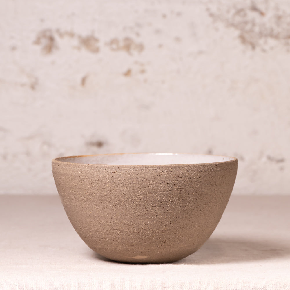 A handmade ceramic bowl with a textured, matte taupe-brown exterior and a smooth, glossy off-white interior, set against a rustic, light beige plaster wall.