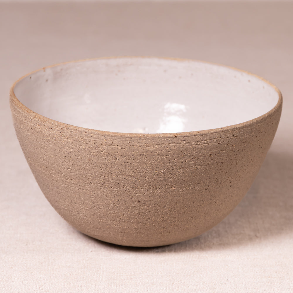 A handmade ceramic bowl with a textured, matte taupe-brown exterior and a smooth, glossy off-white interior, set against a neutral beige background.
