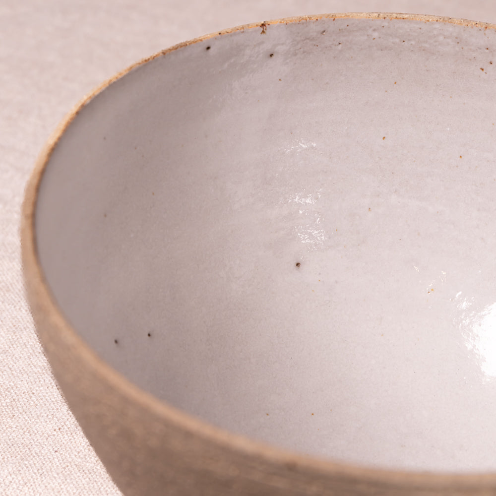 Close-up of a handmade ceramic bowl with a glossy off-white glazed interior and a textured, speckled beige clay rim, set against a neutral beige fabric background.