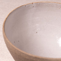 Close-up of a handmade ceramic bowl with a glossy off-white glazed interior and a textured, speckled beige clay rim, set against a neutral beige fabric background.