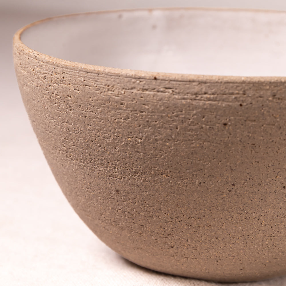 Close-up of a handmade ceramic bowl with a textured, earthy brown exterior and a smooth off-white interior, set against a neutral light background.