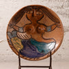 A round handmade ceramic plate featuring a stylized female figure with a face, long hair, and blue and green garments, displayed on a metal stand against a textured white wall.