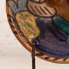 Close-up of a hand-painted ceramic plate or decorative tile featuring a stylized figure with blue, teal, tan, and green glazes on a brown base, mounted on a small stand.