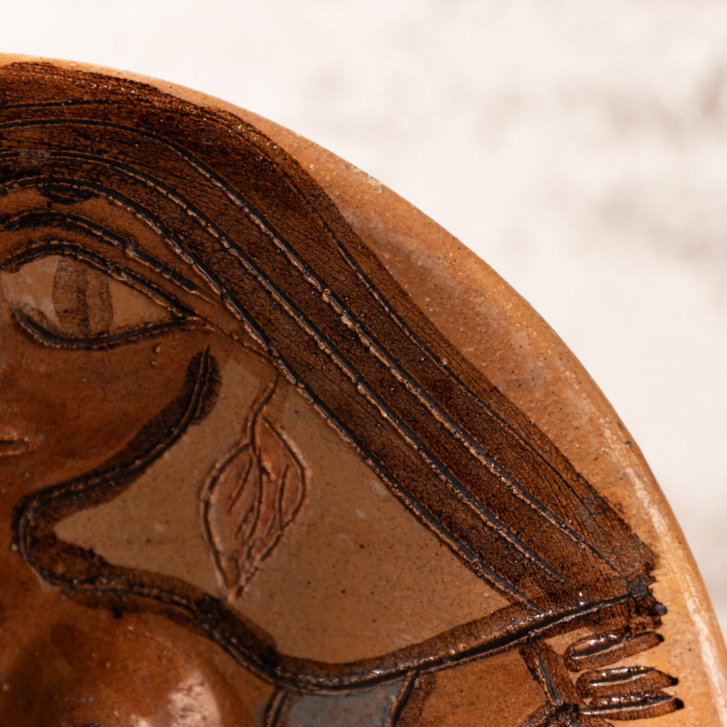 Close-up detail of a terracotta-colored ceramic plate or dish featuring a stylized face with dark brown painted hair and facial features, set against a light neutral background.