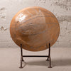 A large round terracotta-colored decorative sculpture or wall plaque with a glossy finish and abstract painted lines, displayed on a dark metal stand against a textured light gray wall.