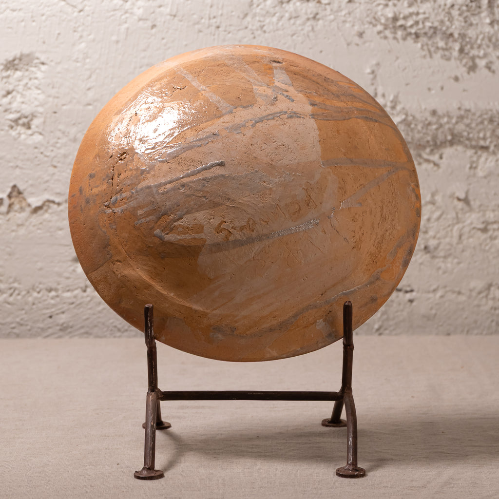 A large round terracotta-colored decorative sculpture or wall plaque with a glossy finish and abstract painted lines, displayed on a dark metal stand against a textured light gray wall.