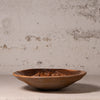 A rustic hand-carved wooden bowl with a natural organic pattern, displayed on a neutral surface against a textured plaster wall.