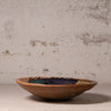 A handmade ceramic shallow bowl with a rustic terracotta base and a glazed interior featuring dark blue, green, and gold accents, set against a textured plaster wall.