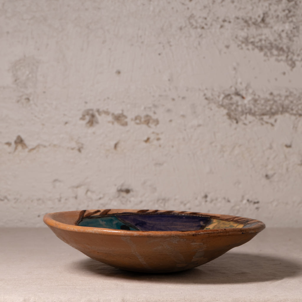 A handmade ceramic shallow bowl with a rustic terracotta base and a glazed interior featuring dark blue, green, and gold accents, set against a textured plaster wall.