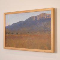 A framed landscape painting of a mountain range over a grassy field, shown hanging on a light beige wall. The artwork features a light wood frame.