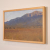 A framed landscape painting of a mountain range over a grassy field, shown hanging on a light beige wall. The artwork features a light wood frame.