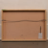 The back of a framed art print or canvas, showing a light natural wood frame, a brown cardboard backing, a hanging wire, and a small white tag, set against a textured off-white wall and dark wooden floor.