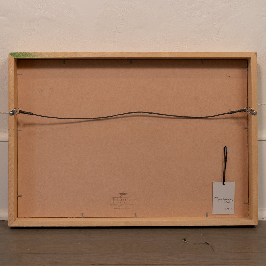 The back of a framed art print or canvas, showing a light natural wood frame, a brown cardboard backing, a hanging wire, and a small white tag, set against a textured off-white wall and dark wooden floor.