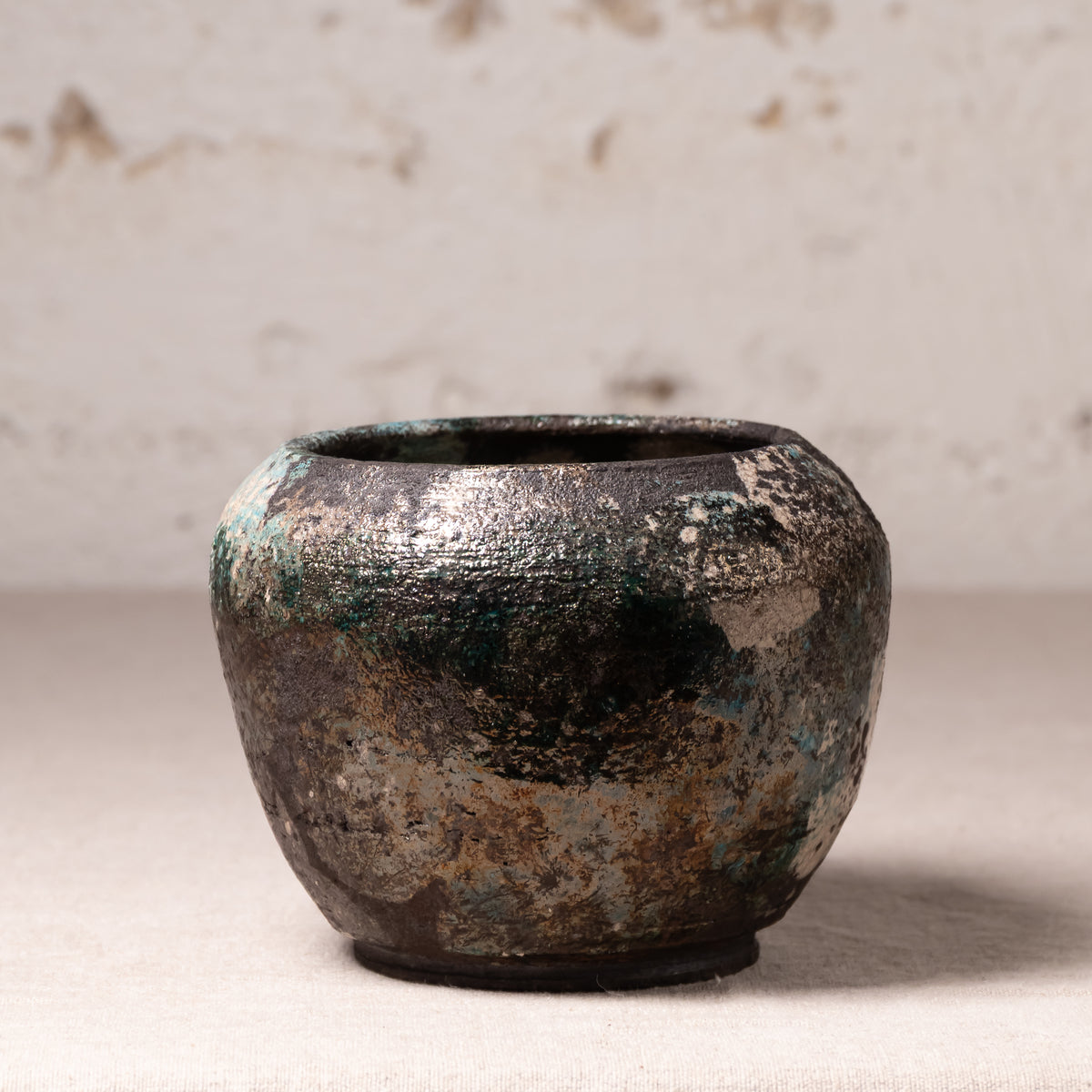 A small handmade ceramic pot with a textured, distressed finish showing dark brown, black, and green patina-like variations, set against a neutral, light beige textured background.