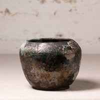 A small handmade ceramic pot with a textured, distressed finish showing dark brown, black, and green patina-like variations, set against a neutral, light beige textured background.
