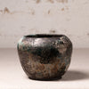 A small handmade ceramic pot with a textured, distressed finish showing dark brown, black, and green patina-like variations, set against a neutral, light beige textured background.