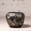A small handmade ceramic pot with a distressed, textured finish featuring a mottled pattern of dark brown, teal, and beige glaze, set against a neutral, light textured background.