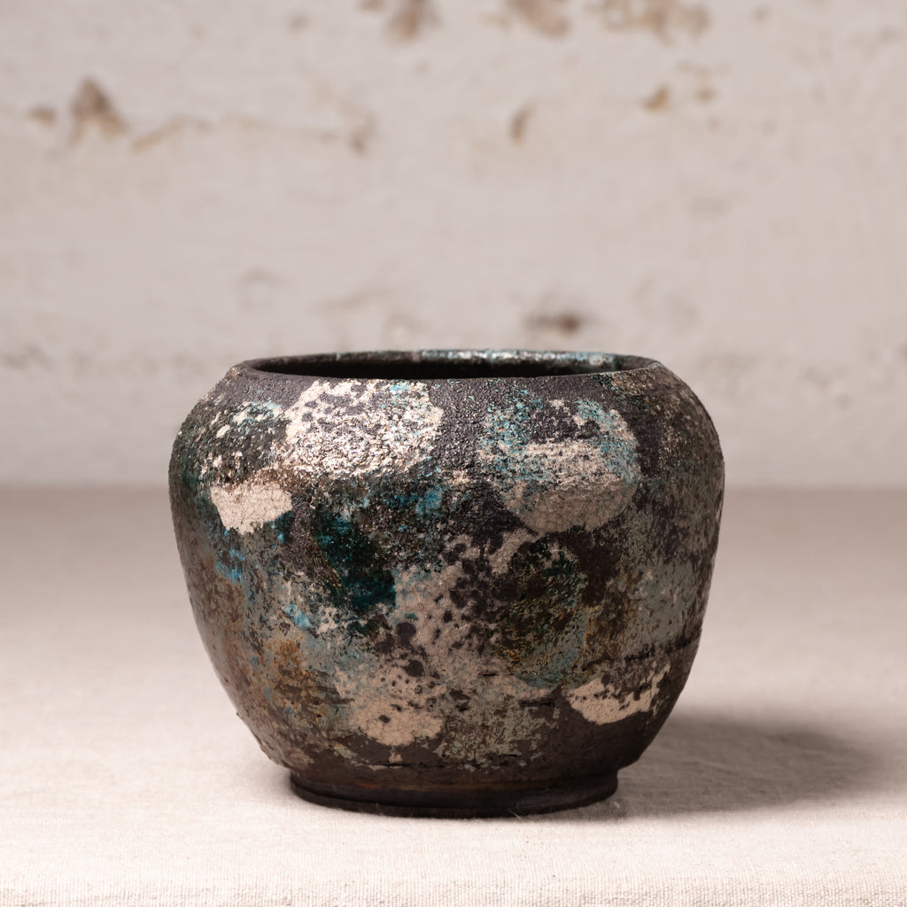A small handmade ceramic pot with a distressed, textured finish featuring a mottled pattern of dark brown, teal, and beige glaze, set against a neutral, light textured background.