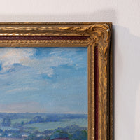 Close-up of an ornate gold picture frame showing part of a landscape painting with blue sky and green fields, mounted on a white wall.