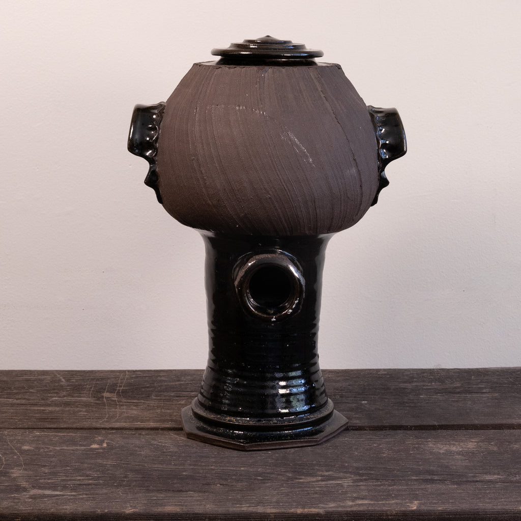A handmade ceramic vessel with a rounded, bulbous body, a glossy black base, and side handles, sitting on a dark wooden surface against a plain light background.