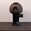A dark brown and black glazed ceramic vessel with a rounded body, a small side protrusion, and a lid, displayed on a dark wooden surface against a plain light wall.