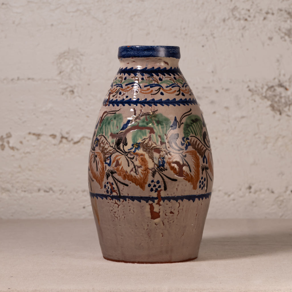 A vintage ceramic vase with a bulbous shape, featuring a hand-painted floral and bird design in blue, green, and brown on a beige stoneware base, set against a textured off-white wall.